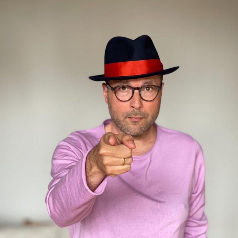 Mann mit Brille und roter Kappe, zeigt mit dem Finger und trägt ein pinkes Oberteil.